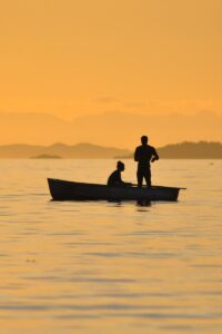 Two people fishing on a calm lake at sunset, silhouetted by the golden sky.